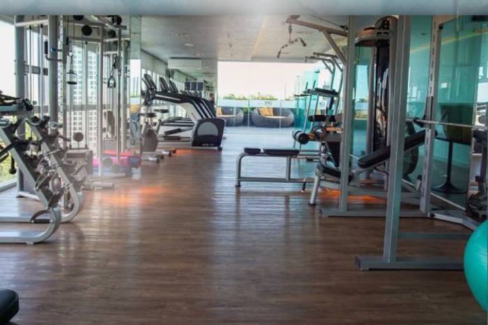 One Tower Pratumnak indoor gym with various exercise machines, wooden floor, and large windows to an outdoor lounge.