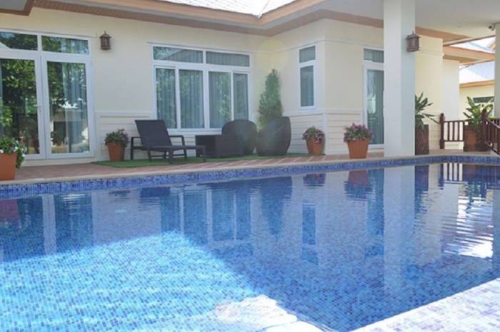Amorn Village house exterior with a swimming pool reflecting the sky, a patio with seating, and large windows.